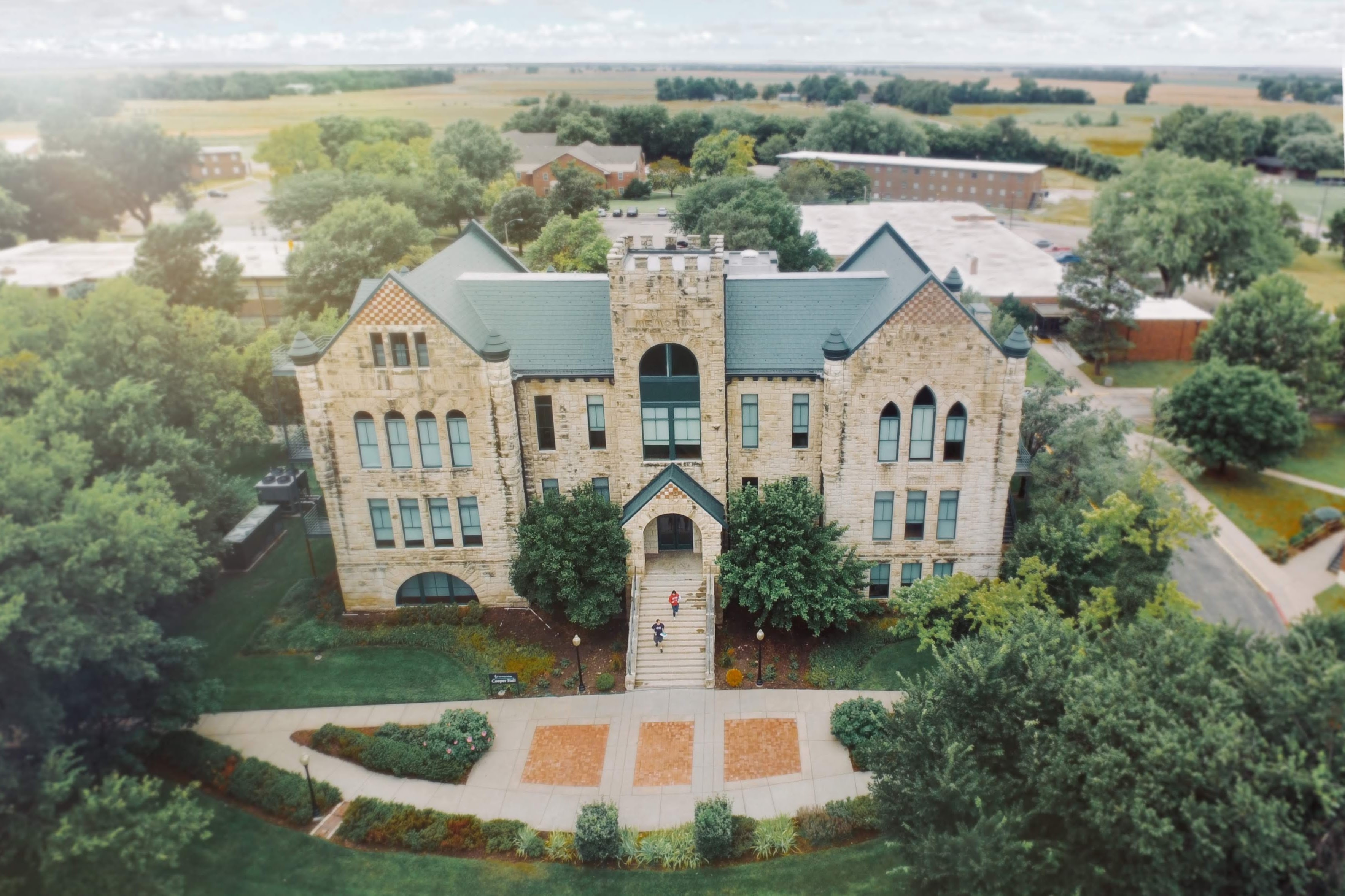 Cooper Hall from the air. The limestone building has a green roof and a square central tower, and stone steps drop from its arched entrance to a paved plaza. Flat Kansas farmland runs out to the horizon behind it.