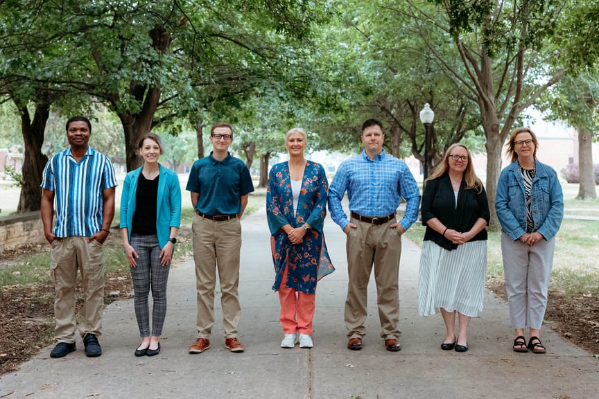 Seven people dressed nicely and standing on a sidewalk with trees behind them