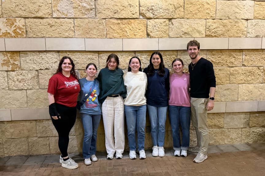 Seven people dressed nicely and standing on a sidewalk with a stone wall or building behind them